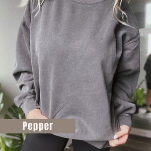 May include: A gray sweatshirt with the word "Pepper" printed on a rectangular label. The long-sleeved top has a crew neck and ribbed cuffs. The garment is a solid color and appears to be made of a soft material.