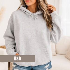 Op de afbeelding: Een lichtgrijze hoodie met een trekkoord in de capuchon en een kangoeroezak aan de voorkant. Het woord "Ash" staat op een bruin rechthoekig label onderaan de afbeelding. De hoodie is gemaakt van een zacht, comfortabel materiaal.