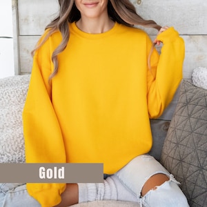 以下が含まれることがあります： 長袖のゴールド色のスウェットシャツ。画像の下部にある茶色の長方形のラベルに「Gold」の文字がプリントされています。スウェットシャツは柔らかい素材でできており、人が着用しています。
