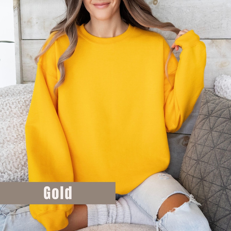 以下が含まれることがあります： 長袖のゴールド色のスウェットシャツ。画像の下部にある茶色の長方形のラベルに「Gold」の文字がプリントされています。スウェットシャツは柔らかい素材でできており、人が着用しています。