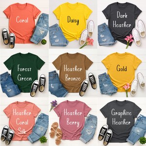 May include: A collection of nine t-shirts in various colors, each paired with denim shorts and sneakers or sandals. The shirts feature text labels like "Coral," "Daisy," "Dark Heather," "Forest Green," "Heather Bronze," "Gold," "Heather Coral Silk," "Heather Berry," and "Graphite Heather."