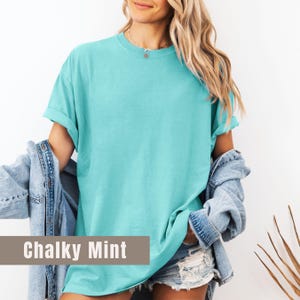 May include: A turquoise, short-sleeved t-shirt with rolled-up sleeves. The shirt is a solid color and has a relaxed fit. The text "Chalky Mint" is displayed on the bottom of the image. A denim jacket is draped over the shoulders.