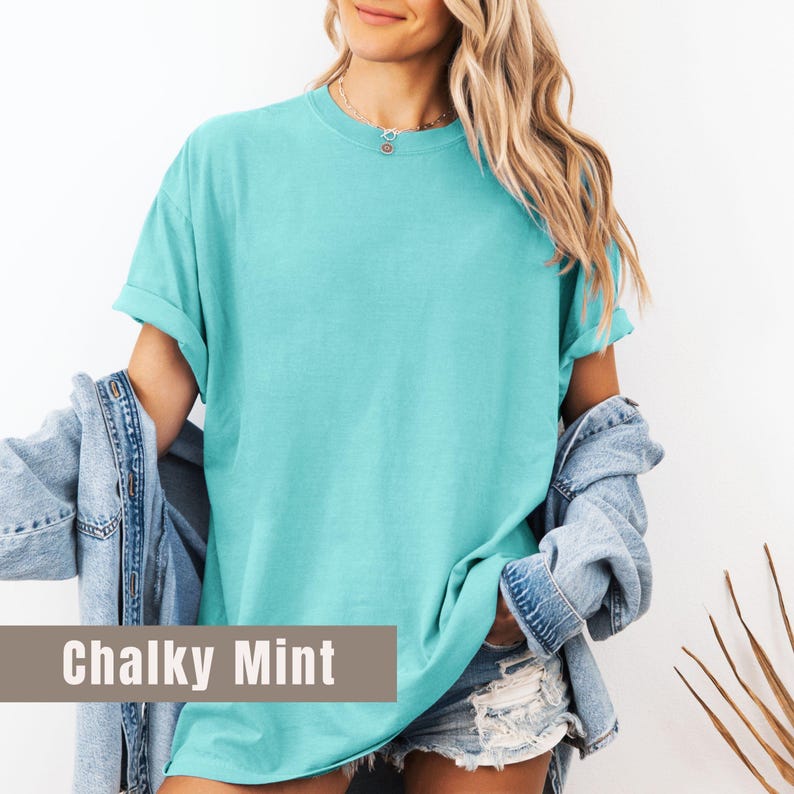 May include: A turquoise, short-sleeved t-shirt with rolled-up sleeves. The shirt is a solid color and has a relaxed fit. The text "Chalky Mint" is displayed on the bottom of the image. A denim jacket is draped over the shoulders.