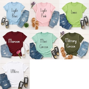May include: A collection of t-shirts in various colors, including light blue, pink, lime, maroon, mint green, military green, and white. Each shirt is paired with denim shorts or jeans and sneakers or sandals. The text on the shirts indicates the color.
