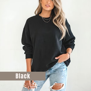 以下が含まれることがあります： 黒いクルーネックのスウェットシャツ。「Black」の文字が、画像の下部にある茶色の長方形のバナーに印刷されています。スウェットシャツは、明るい青色のリップドジーンズと合わせて着用されています。