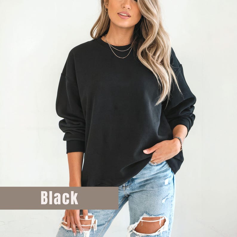 以下が含まれることがあります： 黒いクルーネックのスウェットシャツ。「Black」の文字が、画像の下部にある茶色の長方形のバナーに印刷されています。スウェットシャツは、明るい青色のリップドジーンズと合わせて着用されています。