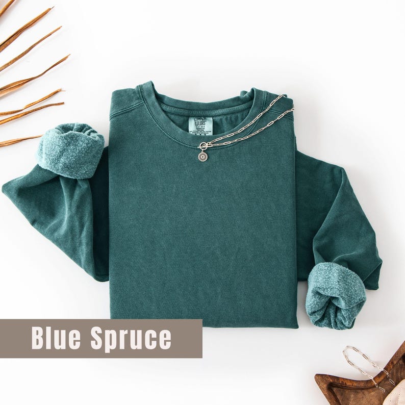 May include: A teal-colored long-sleeve sweatshirt with rolled-up cuffs. A silver chain necklace with a small pendant rests on the shirt. The text "Blue Spruce" is displayed on a brown rectangular background.