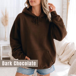 Op de afbeelding: Een donkerchocoladekleurige hoodie met een capuchon met trekkoord en een kangoeroezak. De tekst "Dark Chocolate" wordt weergegeven in een rechthoekige doos onderaan de afbeelding. De hoodie is gemaakt van een zacht, comfortabel materiaal.