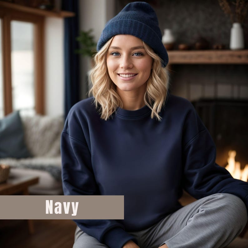 以下が含まれることがあります： ネイビーブルーのビーニーとスウェットシャツを着た人。画像の下部にあるベージュの長方形に「Navy」という文字が表示されています。その人は笑顔で、暖炉の前に座っています。
