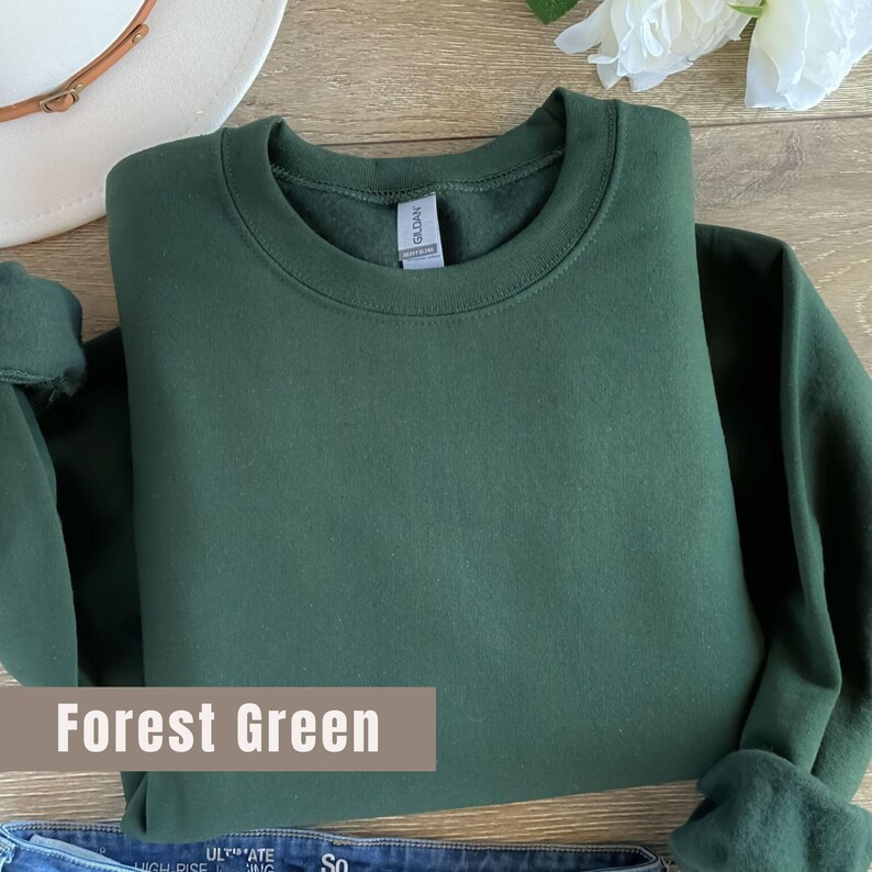以下が含まれることがあります： クルーネック、折り畳まれた袖、タグに「Gildan」のブランド名が入ったフォレストグリーンのスウェットシャツ。「Forest Green」の文字が下部の長方形のボックスに表示されています。白い帽子と花が背景にあります。