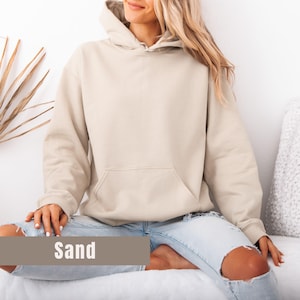 Op de afbeelding: Een zandkleurige hoodie met een voorzak en een capuchon. Het woord "Sand" wordt weergegeven in een bruine rechthoek onderaan de afbeelding. De hoodie wordt gedragen met lichtblauwe jeans.