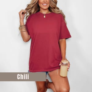 May include: A short-sleeved, oversized t-shirt in a chili red color. The shirt has a crew neck and a relaxed fit. The word "Chili" is displayed on a rectangular label at the bottom of the image. The model is holding a glass with a straw.