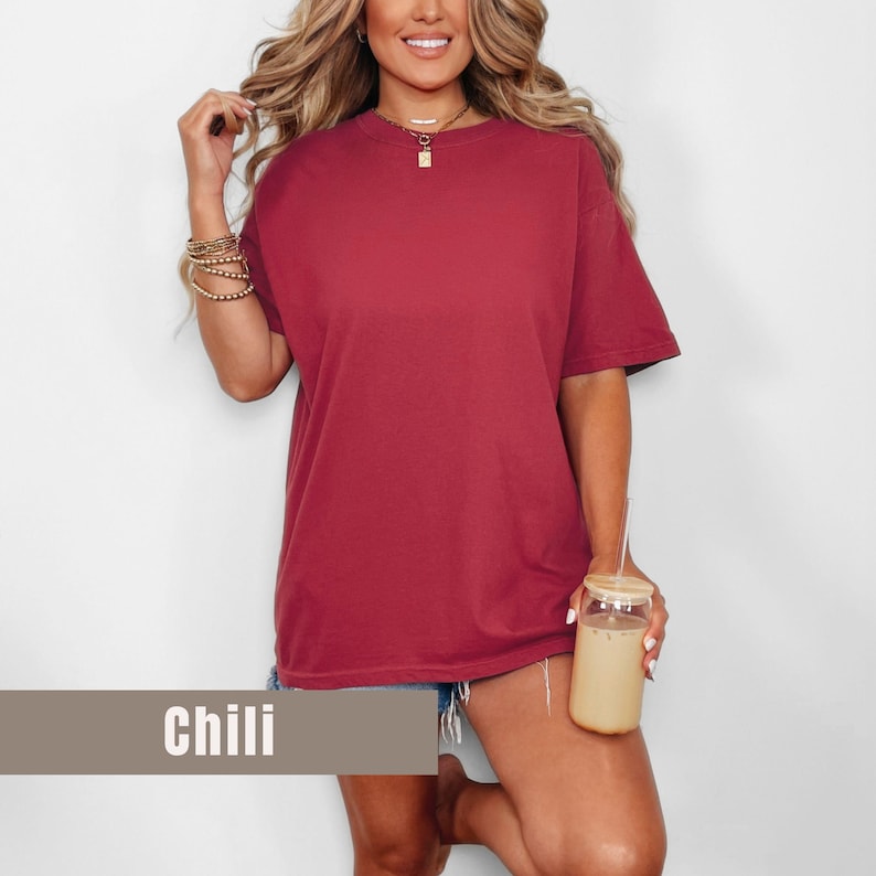 May include: A short-sleeved, oversized t-shirt in a chili red color. The shirt has a crew neck and a relaxed fit. The word "Chili" is displayed on a rectangular label at the bottom of the image. The model is holding a glass with a straw.