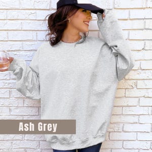 以下が含まれることがあります： アッシュグレーのクルーネックトレーナー。長袖トップスは明るいグレー色です。「Ash Grey」の文字が画像の下部にある茶色の長方形に表示されています。スウェットシャツはカジュアルなスタイルです。
