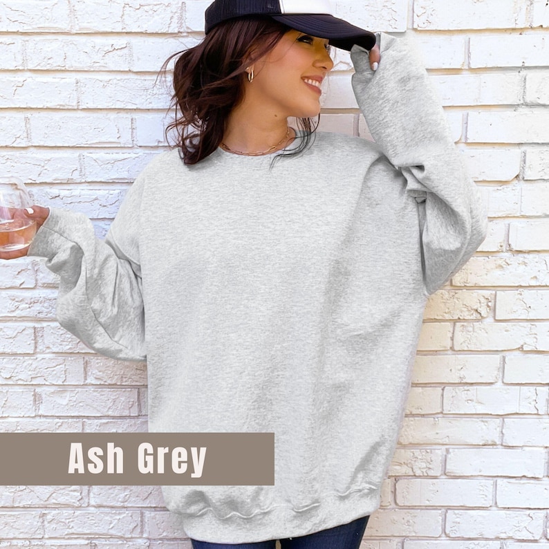 以下が含まれることがあります： アッシュグレーのクルーネックトレーナー。長袖トップスは明るいグレー色です。「Ash Grey」の文字が画像の下部にある茶色の長方形に表示されています。スウェットシャツはカジュアルなスタイルです。