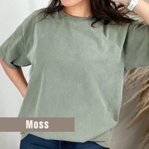 May include: A short-sleeved, moss-colored t-shirt. The shirt has a crew neck and a relaxed fit. The word "Moss" is printed in white on a brown rectangular label at the bottom of the shirt.