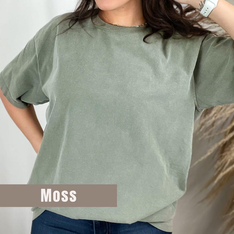 May include: A short-sleeved, moss-colored t-shirt. The shirt has a crew neck and a relaxed fit. The word "Moss" is printed in white on a brown rectangular label at the bottom of the shirt.