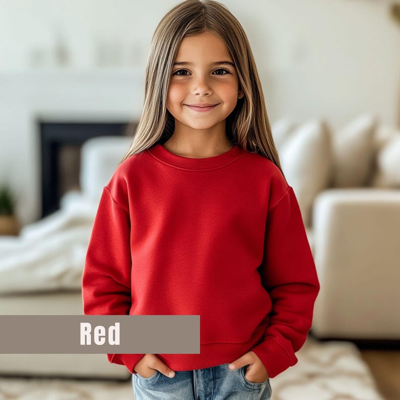 May include: A young person wearing a bright red crew neck sweatshirt. The sweatshirt has long sleeves and a classic, simple design. The word "Red" is displayed in a rectangular box at the bottom of the image. The person is smiling and wearing blue jeans.