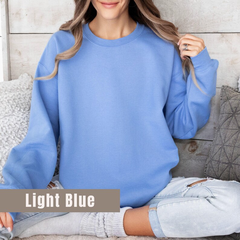 以下が含まれることがあります： クルーネックと長袖のライトブルーのスウェットシャツ。「Light Blue」の文字が、画像の下部にある茶色の長方形のラベルに表示されています。スウェットシャツは柔らかい素材で作られています。