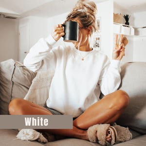 以下が含まれることがあります： 下部に「White」という文字が入ったバナーが付いた、白いクルーネックのスウェットシャツ。その人は黒いマグカップを持っています。その人は金のネックレスとふわふわのスリッパを履いています。