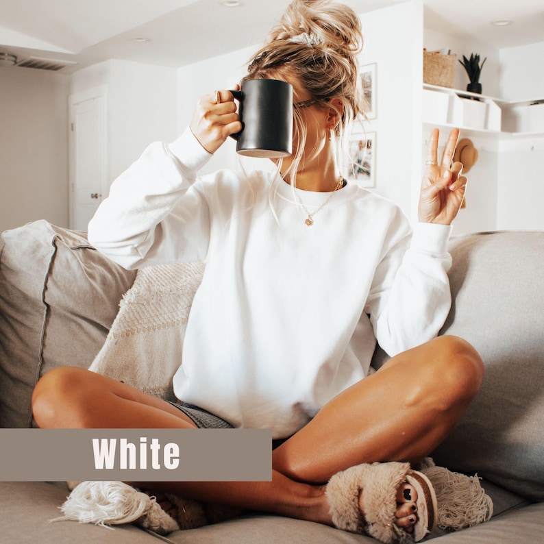 以下が含まれることがあります： 下部に「White」という文字が入ったバナーが付いた、白いクルーネックのスウェットシャツ。その人は黒いマグカップを持っています。その人は金のネックレスとふわふわのスリッパを履いています。