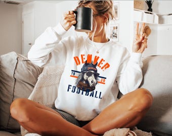 May include: White sweatshirt with a graphic of a horse wearing a beanie and sunglasses, with the text "DENVER FOOTBALL" in orange and navy blue. The sweatshirt has long sleeves.