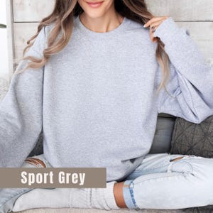 以下が含まれることがあります： 茶色の長方形に「Sport Grey」の文字が入った、ライトグレーのスウェットシャツ。長袖トップスは柔らかい素材で作られています。スウェットシャツはカジュアルなスタイルです。