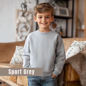 May include: A light gray sweatshirt with the text "Sport Grey" is worn by a smiling child. The sweatshirt has a crew neck and long sleeves. The child is wearing blue jeans.