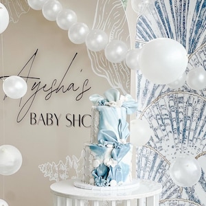 Könnte beinhalten: Eine zweistöckige Babyparty-Torte mit blau-weißer Glasur, verziert mit dekorativen Schleifen und Blumen. Die Torte steht auf einem weißen Tisch, mit Luftballons und einem Hintergrund mit der Aufschrift "Ayesha's BABY SHC".