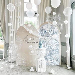 Könnte beinhalten: Eine Babyparty-Szene mit weißen Luftballons, einer Torte und dekorativen Bögen. Die Bögen haben Muschel- und Fächerdesigns. Der Text "Ayla's Baby Shower" ist auf einem Bogen zu sehen. Ein weißer, verzierter Tisch trägt die Torte.