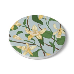 May include: Round coaster featuring a botanical illustration of vanilla flowers and leaves in shades of yellow, green, and brown on a light blue background. The design is a detailed, hand-drawn style.