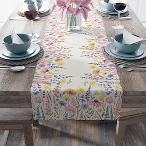 May include: A floral table runner with a watercolor design featuring pink, yellow, and purple flowers. The runner is placed on a wooden table set for a meal, with plates, bowls, and wine glasses.
