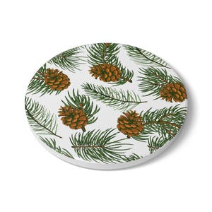 May include: A round, white coaster featuring a repeating pattern of detailed brown pine cones and green pine needles. The design is a hand-drawn style, creating a natural and rustic aesthetic. The coaster is perfect for protecting surfaces from drink rings.