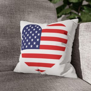 Può includere: Cuscino quadrato bianco con un disegno a forma di cuore della bandiera americana. Il disegno presenta un campo blu con stelle bianche e strisce rosse e bianche. Il cuscino è su un divano grigio.