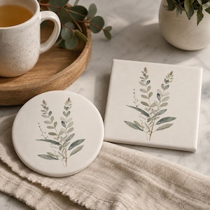 Könnte beinhalten: Ein runder Keramikuntersetzer und ein quadratischer Keramikuntersetzer, beide mit einer cremeweißen Grundfarbe und einer zarten Aquarellillustration von grünen Eukalyptusblättern und Stängeln, die jeweils mittig auf der Oberfläche abgebildet sind.