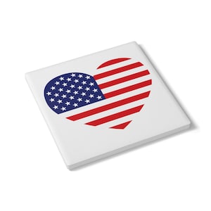 Può includere: Un sottobicchiere quadrato bianco con un disegno a forma di cuore della bandiera americana. Il cuore è composto da strisce rosse e bianche e da una sezione blu con stelle bianche, che rappresenta gli Stati Uniti d'America.