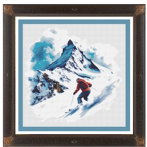 May include: Framed cross-stitch art featuring a skier descending a snowy mountain. The artwork is set against a blue sky and includes the text "Hitting The Slopes" at the top. The frame is dark brown.