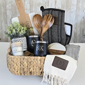 May include: A wicker basket filled with a housewarming gift set. The set includes a wooden cutting board, two wooden spoons, a black oven mitt, two black and white coasters, a white candle, a black and white patterned planter with a small green plant, two black insulated tumblers with personalized initials, and a white towel with a brown leather patch with a monogram.