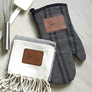 May include: A black and white striped oven mitt and a cream-colored kitchen towel with a herringbone pattern and fringe. Both items feature a brown rectangular patch with the text "home SWEET HOME" printed in black script and block lettering.