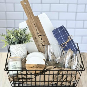 May include: A black wire basket filled with a housewarming gift set. The set includes a wooden and marble cutting board, two wine glasses, a candle, coasters, and a blue kitchen towel. The cutting board and coasters are engraved with the text "clark est. july 2023". The candle has the text "here's to new beginnings".