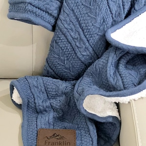 May include: A blue cable-knit blanket with a white sherpa lining. The blanket has a brown leather patch with the name "Franklin" and a mountain graphic. The blanket is folded on a light-colored surface.