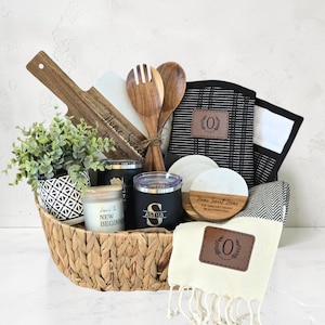 May include: A woven basket containing a wooden cutting board, two wooden salad servers, a black and white patterned planter with a faux plant, two black tumblers, a candle, round coasters, a black and white towel, and a cream-colored fringed towel.