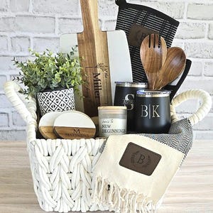 May include: A white woven basket filled with kitchen and home goods. Includes a wooden cutting board, black tumblers, a small plant, coasters, a candle, wooden utensils, a towel, and oven mitts. The cutting board has the text "Miller family".