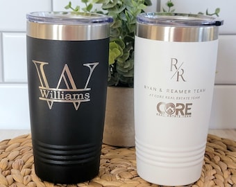 May include: Two insulated tumblers, one black and one white, with silver lids. The black tumbler features the name "Williams" with a stylized "W". The white tumbler displays "RYAN & REAMER TEAM" and "CORE REAL ESTATE TEAM".