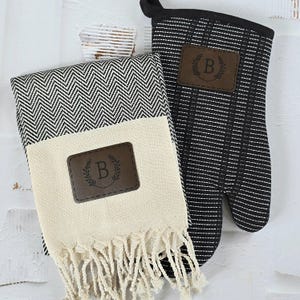 May include: A black and white chevron patterned kitchen towel with a brown leather patch that says "B" in a wreath. A black and white striped oven mitt with a brown leather patch that says "B" in a wreath.