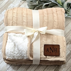 May include: A beige cable knit throw blanket with a white sherpa lining. The blanket is tied with a white satin ribbon and bow. A brown leather patch with the initials "ANH" is sewn onto the blanket.
