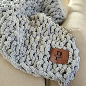 May include: A gray chunky knit throw blanket with a brown leather patch that says "PELLETIER" with a capital "P" in the center.