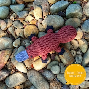 May include: A crocheted plush toy platypus with a red body, blue beak, and black feet. The toy is lying on a bed of rocks. The text "Platypus - Crochet Pattern Only" is in a yellow speech bubble.
