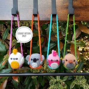 Puede incluir: Cuatro peluches de animales de crochet colgando de ganchos. Los peluches son un pájaro amarillo, un koala gris, un pájaro rosa y un pájaro marrón. Cada peluche está unido a una cuerda de colores.