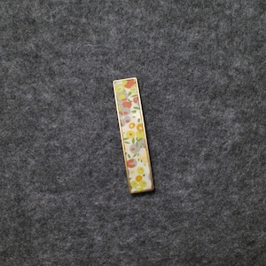 May include: A rectangular tie bar featuring a gold-toned metal frame and a decorative inlay. The inlay displays a repeating pattern of stylized flowers and leaves in shades of orange, yellow, light green, and grey against a white background.